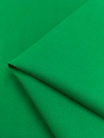 Close-up of a smooth, vividly green piece of folded, medium to heavy weight fabric. The texture appears soft, with fine, even threads creating a uniform surface. The Milano Ponte - Green Tambourine - 165cm from Super Cheap Fabrics is neatly creased, showcasing its flexibility and the richness of its green hue.