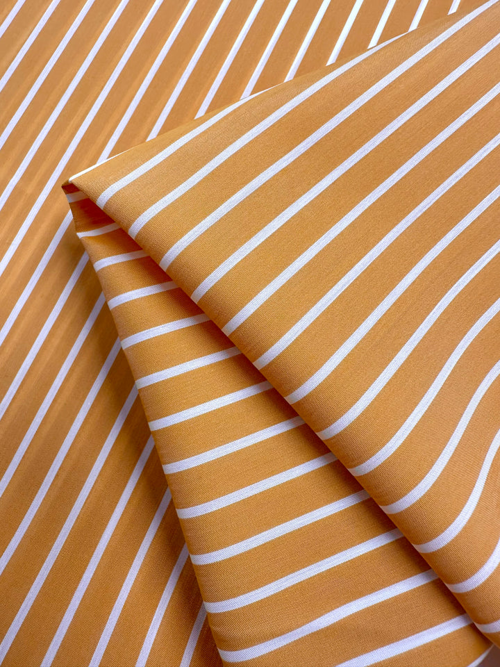 Close-up of Super Cheap Fabrics' Polished Cotton Voile - Sunrise - 155cm, crafted from sustainable deadstock materials and featuring orange and white diagonal stripes. The fabric is folded to display the pattern from various angles, with evenly spaced lines creating a consistent striped design across this eco-friendly textile.