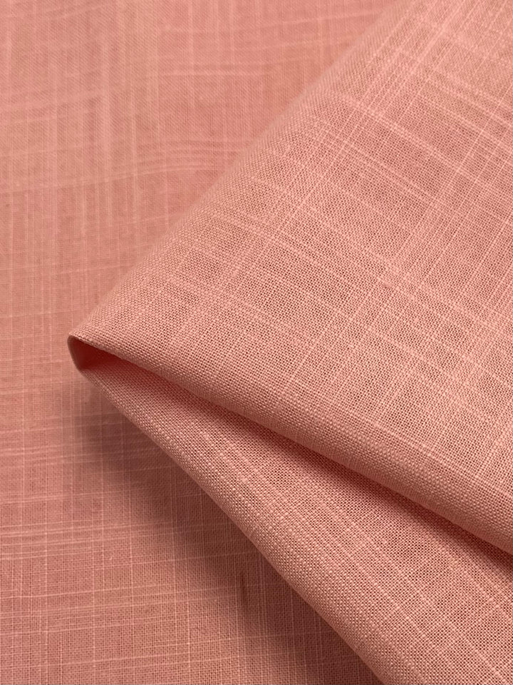 A close-up shot of a folded piece of fabric in a soft, peachy-pink hue. Resembling elegant table linens, the lightweight material showcases a subtle, textured weave with fine lines forming a delicate grid pattern. This is the Linen Blend - Coral Reef - 140cm by Super Cheap Fabrics.
