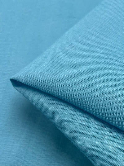 A close-up image of a neatly folded piece of Linen Blend - Norse Blue - 140cm by Super Cheap Fabrics, showcasing its textured surface. The material appears smooth and finely woven, with a clean, crisp fold. The background is the same light blue fabric, creating a harmonious, monochromatic look.