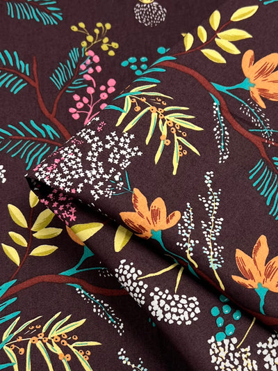 Close-up of a folded piece of Printed Cotton - Botanic Earth - 148cm by Super Cheap Fabrics with a dark burgundy background, featuring a botanical pattern of colorful leaves, flowers, and branches in shades of yellow, pink, green, and turquoise. The design is intricate and vibrant, giving a lively appearance to the lightweight fabric.