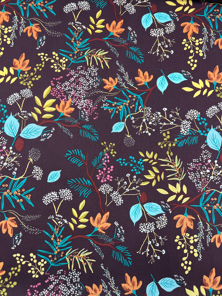 A vibrant botanical pattern features various leaves, berries, and flowers in bright colors like blue, yellow, orange, and white, set against a dark purple background. Printed on lightweight natural cotton fibers by Super Cheap Fabrics, the design of Printed Cotton - Botanic Earth - 148cm is intricate with different shapes and sizes of foliage creating a lively composition.