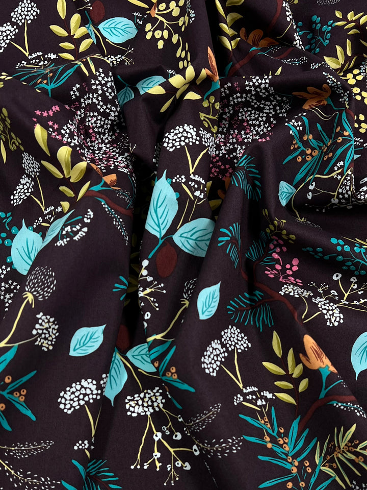 A close-up view of a dark, lightweight fabric featuring a floral pattern. The design includes various flowers, leaves, and branches in shades of blue, white, yellow, and green, creating a colorful, nature-inspired motif. Made from natural cotton fibers, the fabric appears slightly gathered and textured. This is the Printed Cotton – Botanic Earth – 148cm by Super Cheap Fabrics.