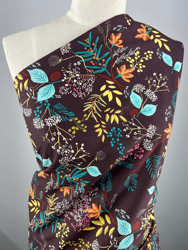 A fabric draped over a mannequin, featuring a dark brown background with a vibrant floral pattern in various colors, including orange, teal, green, yellow, and white. Made from lightweight Printed Cotton - Botanic Earth - 148cm by Super Cheap Fabrics, the design includes flowers, leaves, and small berries for a lively and colorful look.