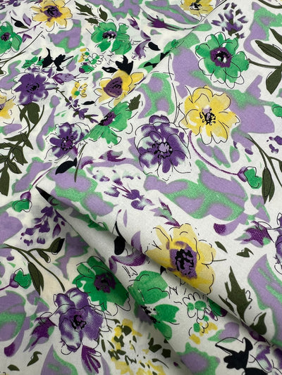 Image of folded, patterned fabric with a floral design. The flowers are in shades of purple, yellow, and green, set against a light background. Made from 100% cotton, the design includes leaves and vines in coordinating colors, creating a detailed, vibrant print. Introducing Printed Cotton - Comic Field - 148cm by Super Cheap Fabrics.