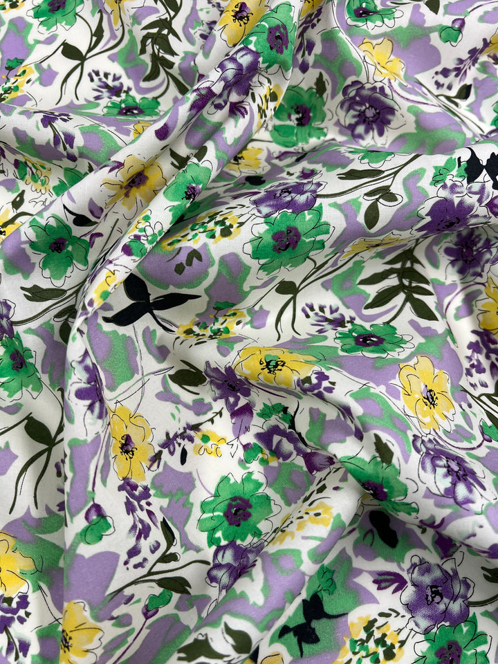 A piece of Super Cheap Fabrics' Printed Cotton - Comic Field - 148cm featuring a vibrant floral print with green, yellow, purple, and black flowers scattered over a white background. Made from 100% cotton, the fabric is gently draped, creating soft folds and shadows that enhance the texture and color contrast of the design.