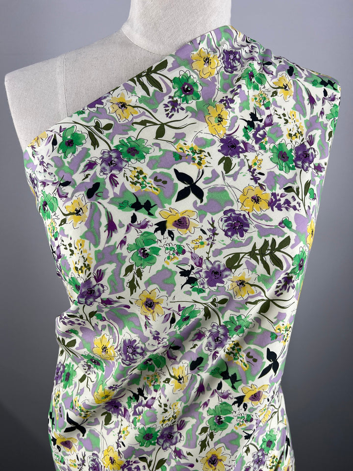 A mannequin draped with Printed Cotton - Comic Field - 148cm, featuring a vibrant floral pattern of green, purple, yellow, and black flowers on a white background. The natural cotton fibers from Super Cheap Fabrics are arranged to create a one-shoulder display against a plain grey backdrop.