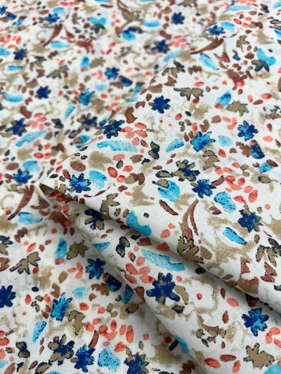 A close-up image of a Printed Cotton - Natures Mark - 148cm fabric with a colorful floral pattern. The design includes blue, coral, and beige flowers with greenish-brown stems and leaves, scattered on a white background. This versatile multiuse textile from Super Cheap Fabrics is made from natural cotton fibers, with part of the fabric slightly folded over itself.