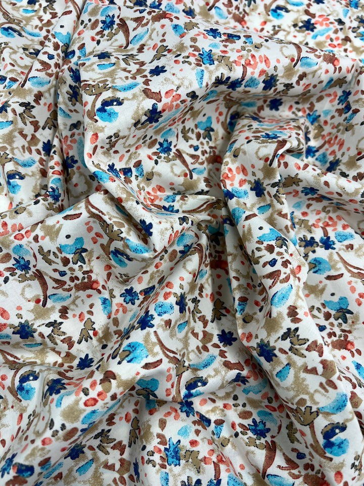 A close-up of a crumpled fabric featuring a floral pattern. The Super Cheap Fabrics Printed Cotton - Natures Mark - 148cm is adorned with blue, brown, and pink flowers set against a cream-colored background. The design includes various leaf and petal shapes, creating a detailed and intricate appearance.