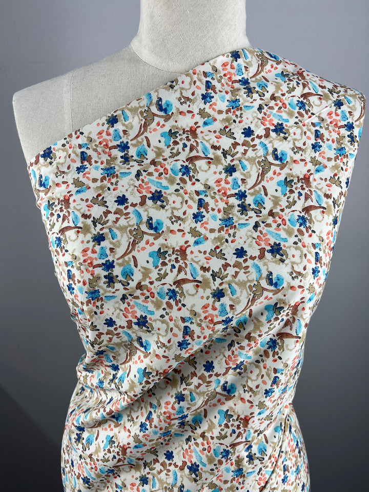 A mannequin draped in a Super Cheap Fabrics Printed Cotton - Natures Mark - 148cm fabric with a colorful floral pattern, featuring blue, pink, and brown flowers and leaves. The fabric covers the mannequin asymmetrically from one shoulder to the opposite hip, against a plain gray background.