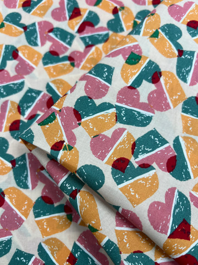 Close-up of **Super Cheap Fabrics Printed Cotton - Fruity Candy Hearts - 148cm** fabric with a colorful pattern featuring hearts segmented into four parts. Each heart segment is filled with varying shades of pink, orange, teal, and small red dots. The natural cotton fibers create a soft texture as the folded fabric displays the pattern evenly.