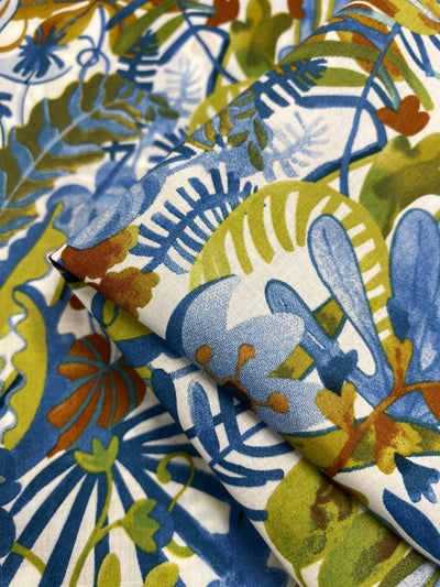 A close-up image of two pieces of Printed Cotton - Aquatic Flora - 148cm by Super Cheap Fabrics with vibrant botanical patterns. The design features leaves, ferns, and abstract plant shapes in blue, green, brown, and orange tones on a white background. One piece of the multi-use textile is slightly overlapping the other.