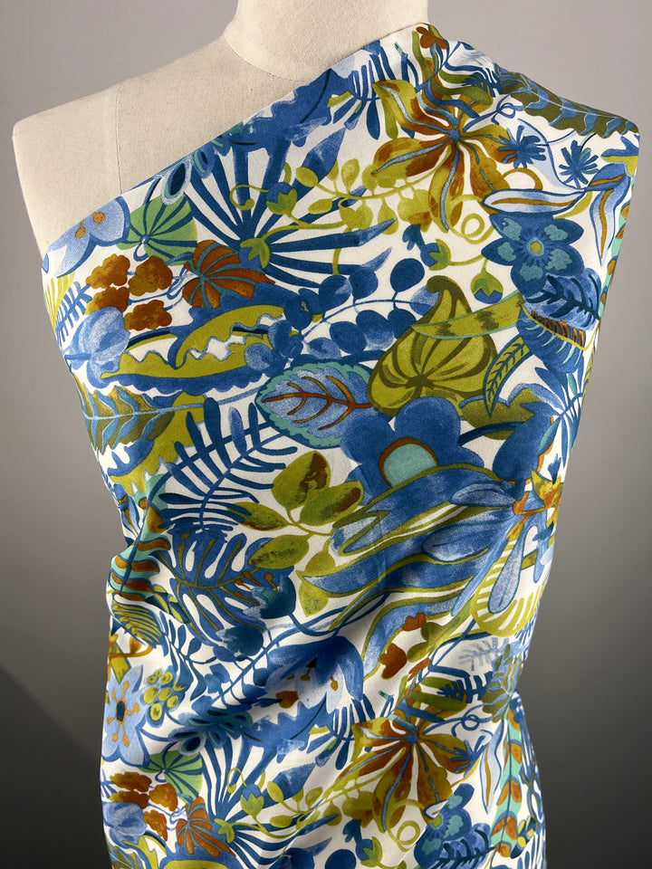 A mannequin is draped in a Super Cheap Fabrics Printed Cotton - Aquatic Flora - 148cm fabric featuring a vibrant, tropical print. The pattern includes various blue, green, yellow, and orange leaves and flowers against a white background. The fabric's intricate and colorful design is showcased beautifully using natural cotton fibers.
