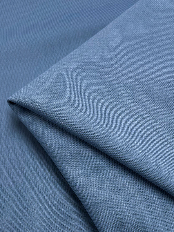 A close-up image of a soft, blue stretch fabric folded neatly. The material has a smooth texture, and the fold creates subtle shadows and highlights, suggesting a gentle and lightweight quality. The fabric depicted is the Ponte - Placid Blue - 155cm from Super Cheap Fabrics.