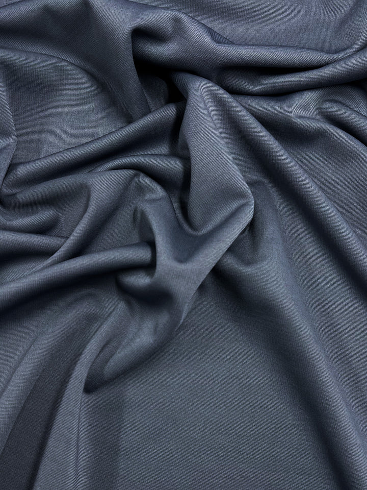 A close-up view of smooth, dark grey stretch fabric with a subtle sheen, draped and crumpled in gentle waves and folds. The material's texture and pattern create soft shadows and highlights. This is Ponte - Grisaille - 155cm by Super Cheap Fabrics.