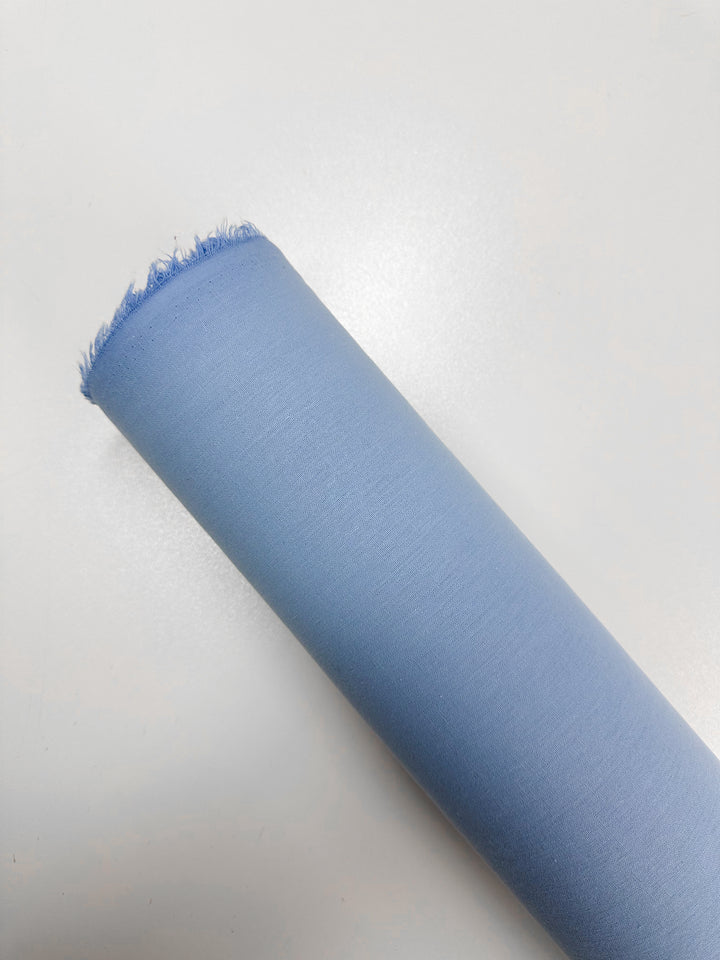 A roll of Ramie Linen - Clear Sky by Super Cheap Fabrics, 150cm wide in light blue with durable natural fibers and frayed edges, sits elegantly on a white background.