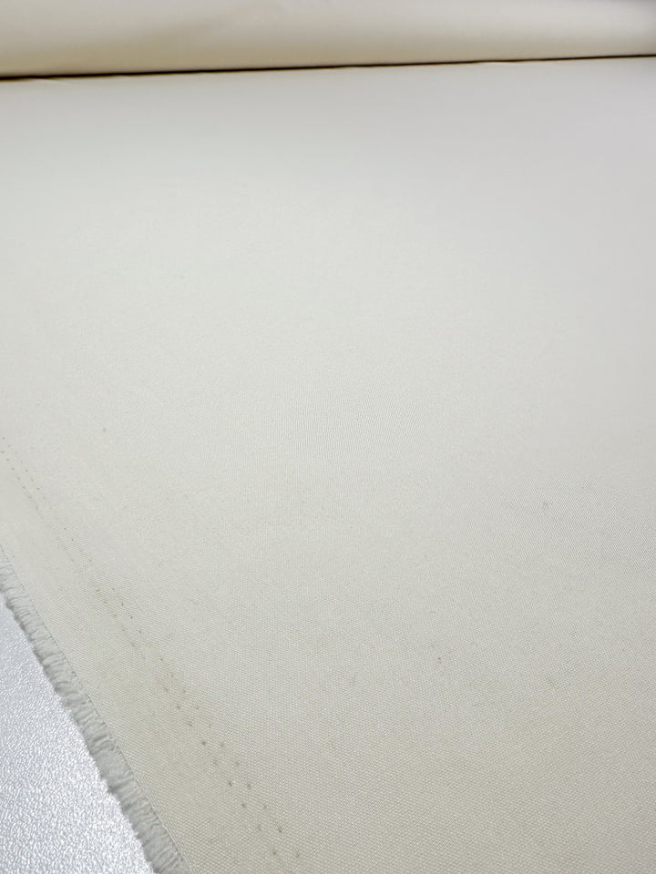 A close-up of Suiting - Pristine - 150cm by Super Cheap Fabrics shows an unrolled, off-white polyester viscose fabric with a slightly textured surface and a frayed edge on the left.
