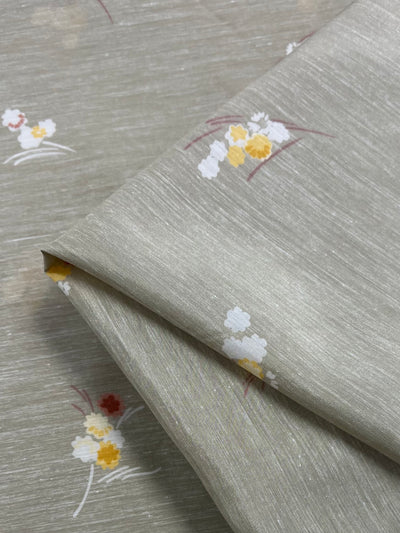 Close-up of a folded piece of Super Cheap Fabrics' Cotton Voile - Little - 120cm with a subtle, light gray textured background. The lightweight fabric features a simple floral design with yellow, white, and red flowers and slender brown branches, perfect for warmer weather.
