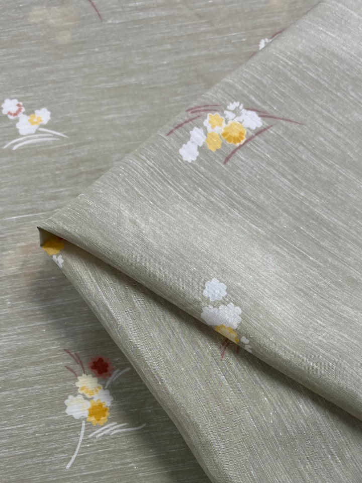Close-up of a folded piece of Super Cheap Fabrics' Cotton Voile - Little - 120cm with a subtle, light gray textured background. The lightweight fabric features a simple floral design with yellow, white, and red flowers and slender brown branches, perfect for warmer weather.