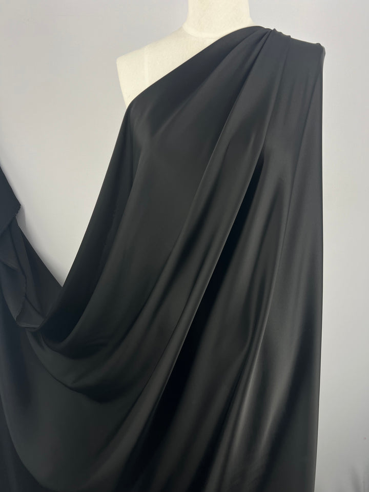 A close-up of a dress form draped in smooth, shiny black satin fabric. The Designer Viscose Satin - Black - 140cm by Super Cheap Fabrics appears to be wrapped around the form, cascading in pleats and folds, showcasing the material's luster and flow.