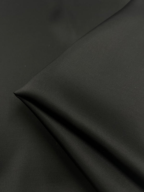 Close-up of Designer Viscose Satin - Black - 140cm from Super Cheap Fabrics with a slight sheen, draped to form a gentle fold or crease. The texture appears soft and the lighting accentuates a subtle shine on the fabric’s surface, making it ideal for lining garments.