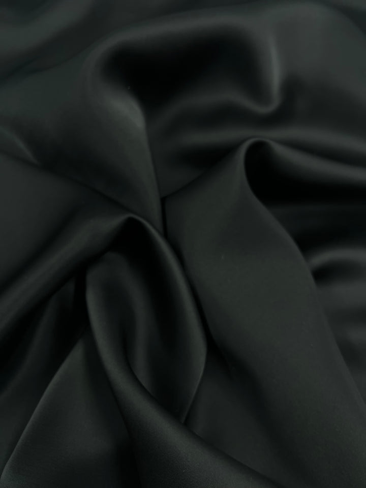 A close-up shot of Designer Viscose Satin - Black - 140cm from Super Cheap Fabrics, showcasing its smooth, glossy texture and folds. The gentle curves and creases capture the luxurious quality of the material, perfect for lining garments.