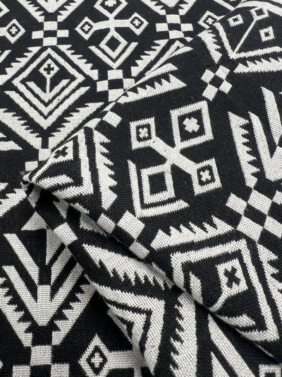 A close-up of folded black fabric featuring intricate white geometric patterns. The design, crafted from 100% Polyester heavy weight fabric, includes a mix of various shapes like diamonds, squares, and triangles, creating a visually striking and symmetrical pattern. This is the Designer Tweed - Mono Aztec - 150cm by Super Cheap Fabrics.