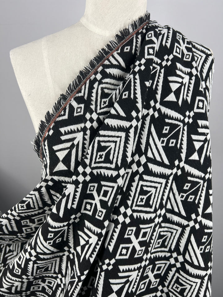 A mannequin draped in Designer Tweed - Mono Aztec - 150cm by Super Cheap Fabrics featuring an intricate black and white geometric pattern. The design includes a variety of shapes such as diamonds, triangles, and squares, giving the appearance of traditional or tribal prints. The 100% polyester fabric has frayed edges.