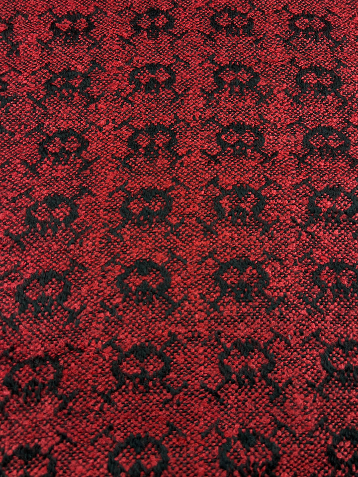 Close-up of Designer Tweed - Lava Serpent - 155cm by Super Cheap Fabrics, a textured red and black heavy weight fabric with an intricate, repeating floral pattern. The design consists of black floral motifs arranged evenly across the vibrant red background. The polyester fabric surface appears woven and has a slightly rough texture.