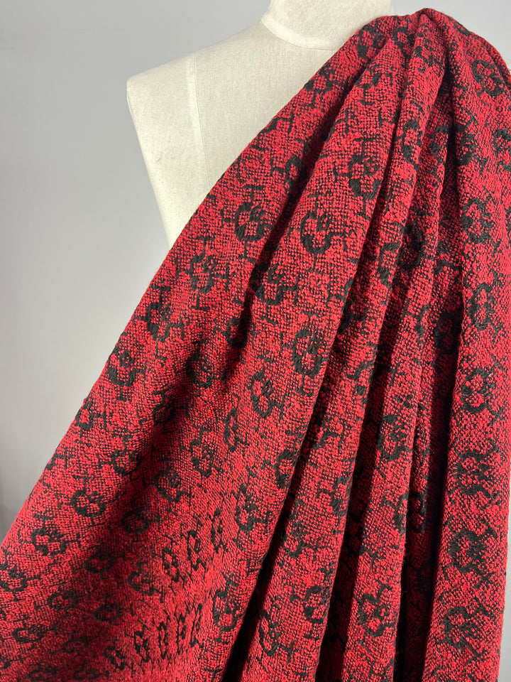 A vibrant red and black textile with an intricate, floral-like pattern draped over a light gray mannequin. The heavy weight fabric appears textured and rich, with its bold design creating a striking contrast against the neutral background. The fabric in question is Designer Tweed - Lava Serpent - 155cm by Super Cheap Fabrics.