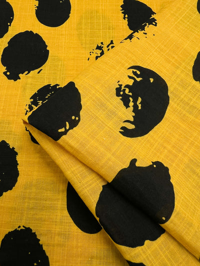 A folded piece of lightweight yellow fabric made of Bamboo Rayon with a pattern of large, irregular black spots. The texture is slightly visible, giving it a woven appearance. The spots vary in size and shape, creating a bold, abstract design perfect for versatile uses. This is the Bamboo Rayon - Stamp Day Lily - 145cm from Super Cheap Fabrics.