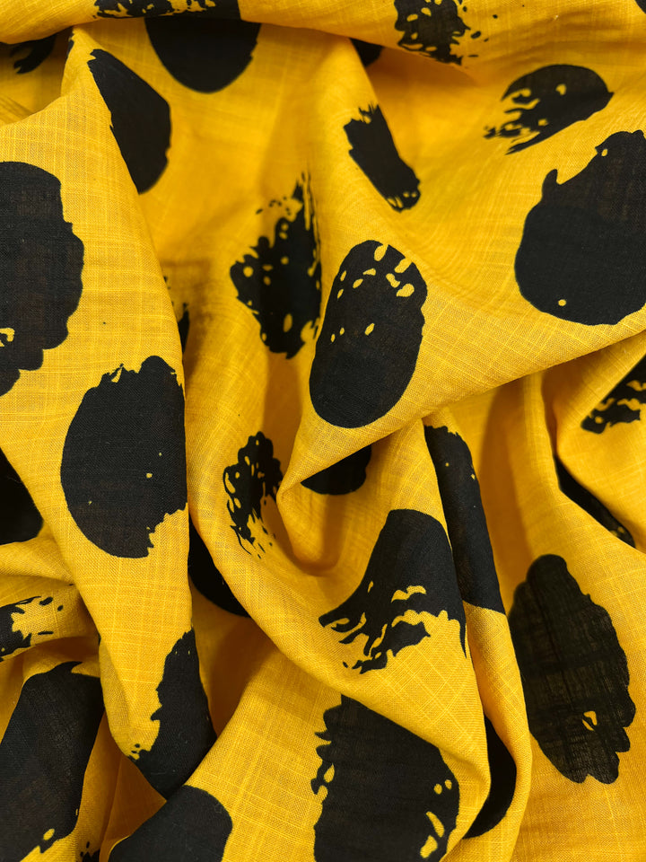 A close-up of Super Cheap Fabrics' Bamboo Rayon - Stamp Day Lily - 145cm with a black abstract circular pattern. The bamboo rayon material is wrinkled and folded, creating a textured appearance with shadows and highlights. The pattern consists of irregularly shaped black spots scattered across the bright yellow background.