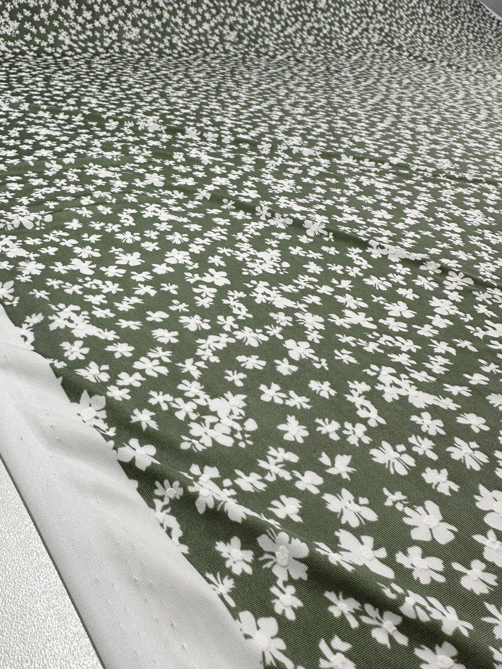 A close-up of Super Cheap Fabrics' Textured Lycra - Verdotone (150cm), a green polyester spandex fabric with a scattered white floral pattern, shown laid out flat with an unfinished white edge on the left.