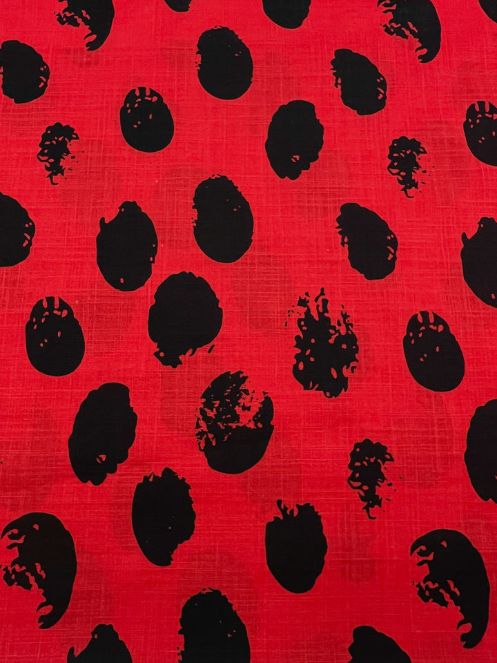 A red lightweight fabric with an abstract pattern of irregular black spots evenly scattered across the surface. The black spots vary in size and have a textured, somewhat frayed appearance, making it ideal for stylish home decor projects. This is the Bamboo Rayon - Stamp High Risk Red - 145cm from Super Cheap Fabrics.