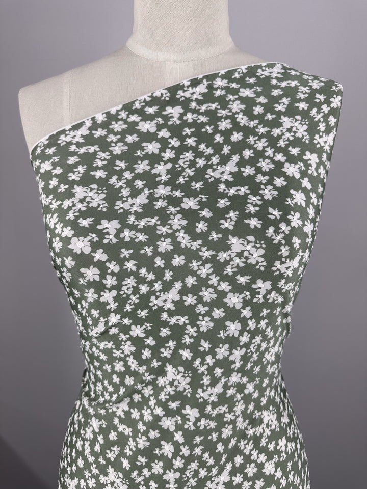 A dress form displays Super Cheap Fabrics' Textured Lycra - Verdotone (150cm), a green printed lycra with a white floral pattern, draped asymmetrically across one shoulder against a plain gray background.