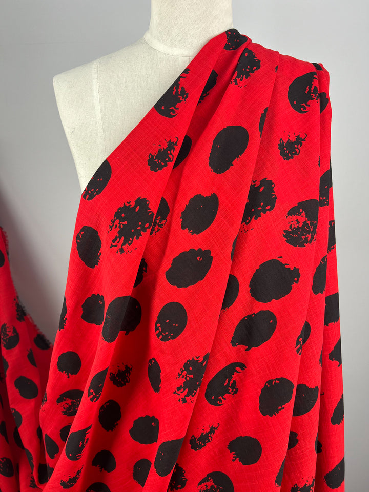 A mannequin draped with a vibrant red lightweight fabric featuring an abstract black polka dot pattern. The Super Cheap Fabrics Bamboo Rayon - Stamp High Risk Red - 145cm material flows smoothly over the mannequin's shoulder against a plain gray background, adding an elegant touch to any home decor.