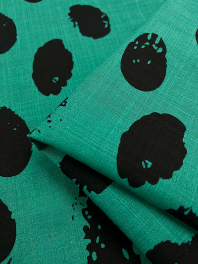 Close-up of a green Bamboo Rayon - Stamp Deep Green - 145cm fabric from Super Cheap Fabrics with large irregular black spots. The fabric has a slight fold, creating a layered effect. Perfect for home decor, the texture appears to be lightweight and slightly textured.