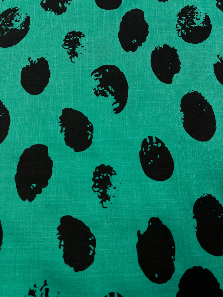 A green **Super Cheap Fabrics Bamboo Rayon - Stamp Deep Green - 145cm** featuring a pattern of irregular black ovals and circular shapes randomly scattered across the surface. Some shapes have texture and appear slightly worn or distressed, creating a dynamic and abstract design, ideal for stylish home decor.