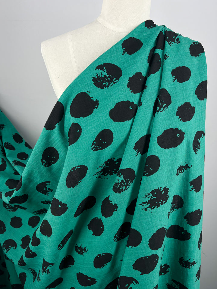 A white mannequin is draped with vibrant teal Bamboo Rayon - Stamp Deep Green - 145cm by Super Cheap Fabrics, featuring a pattern of large, irregular black spots. The lightweight fabric texture appears soft and airy, creating a casual and artistic look.