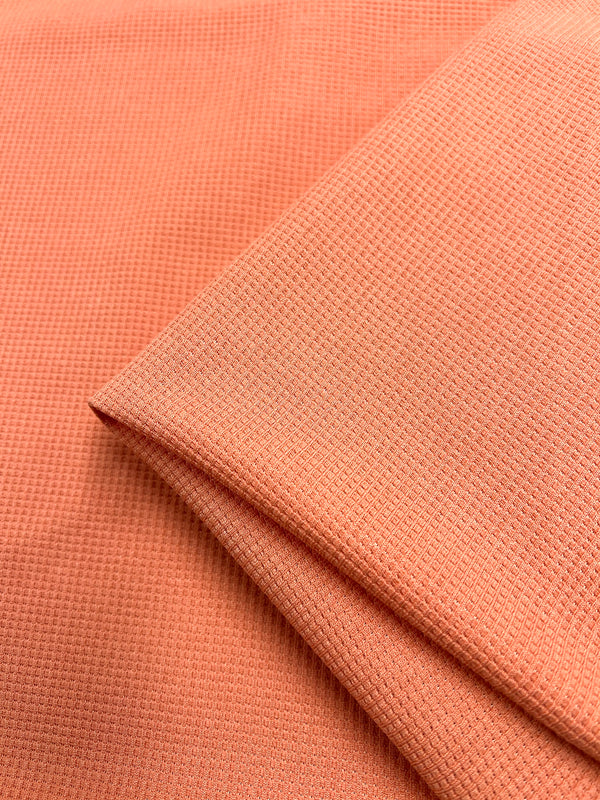 Close-up image of a folded piece of textured orange fabric with a honeycomb weave pattern. The three-dimensional fabric appears smooth and neatly arranged, showcasing its surface detail and vibrant color. Waffle Knit - Blooming Dahlia - 170cm by Super Cheap Fabrics.