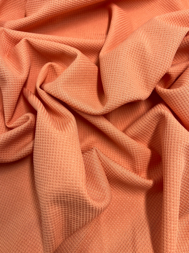 Close-up of a crumpled, textured fabric in a bright orange hue. The Waffle Knit - Blooming Dahlia - 170cm from Super Cheap Fabrics has a small, rectangular grid pattern and soft folds that create depth and shadows. The material appears to be lightweight and breathable.