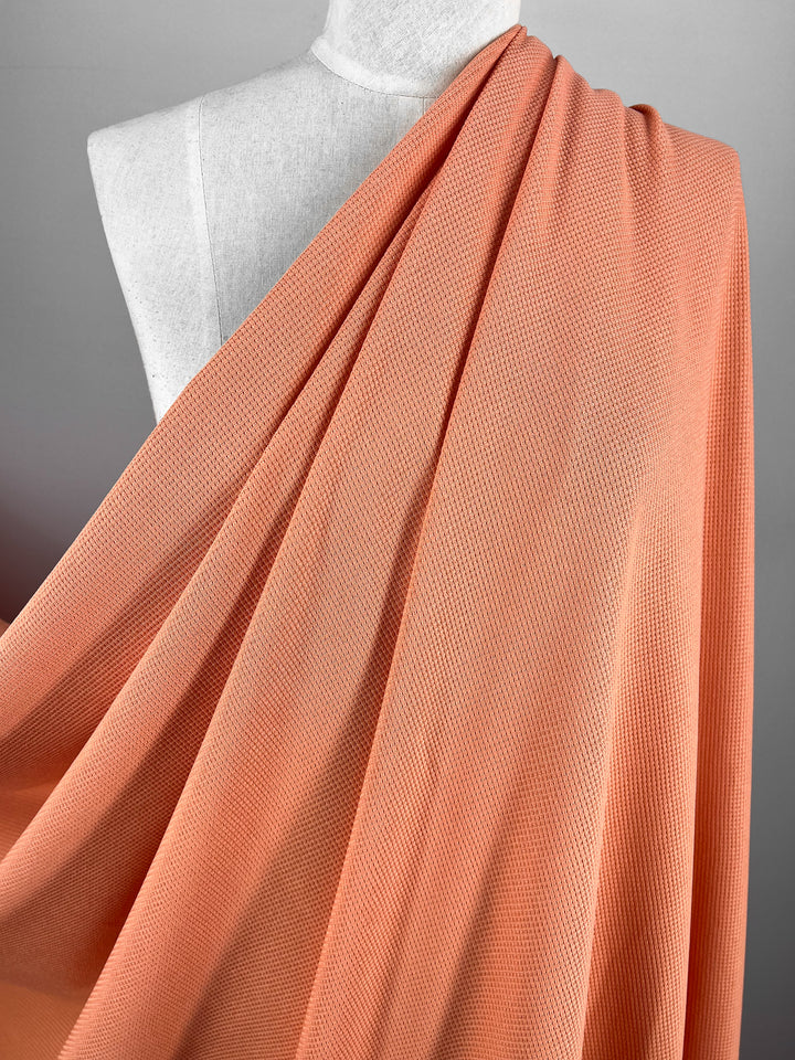 A fabric named Waffle Knit - Blooming Dahlia - 170cm from Super Cheap Fabrics draped elegantly over a white dress form on a neutral gray background. The textured material cascades smoothly, revealing its subtle pattern and soft folds.
