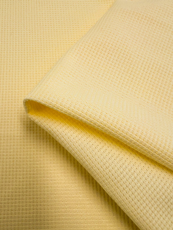 Close-up of a folded, yellow Waffle Knit - Pale Banana - 170cm from Super Cheap Fabrics. The Waffle Knit - Pale Banana's texture, resembling honeycomb fabric, includes small, repeating grid patterns characteristic of waffle-weave fabric. The image highlights the three-dimensional effect and soft, cozy material.