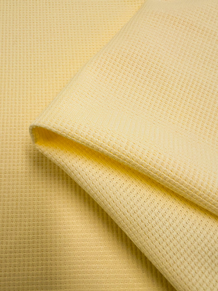 Close-up of a folded, yellow Waffle Knit - Pale Banana - 170cm from Super Cheap Fabrics. The Waffle Knit - Pale Banana's texture, resembling honeycomb fabric, includes small, repeating grid patterns characteristic of waffle-weave fabric. The image highlights the three-dimensional effect and soft, cozy material.