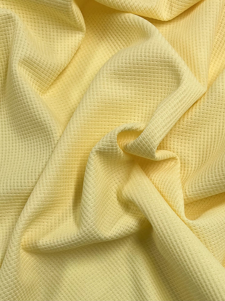 A close-up view of a textured, yellow Waffle Knit - Pale Banana - 170cm fabric by Super Cheap Fabrics, displaying soft folds and creases. The material has a honeycomb pattern, adding depth and dimension to the surface. The light hue and texture create a warm and cozy appearance with a subtle three-dimensional effect.