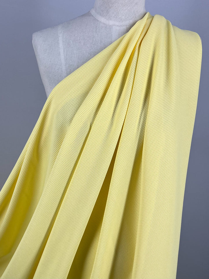 A mannequin draped in a textured, pale yellow waffle fabric is seen against a plain background. The fabric, Waffle Knit - Pale Banana - 170cm by Super Cheap Fabrics, with its subtle weave pattern, creates a three-dimensional effect as it flows smoothly over the mannequin, forming gentle, natural folds.