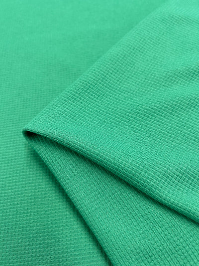 A close-up of an emerald green, honeycomb fabric with a folded corner, revealing its fine grid-like pattern and smooth surface. The fabric appears lightweight and soft, suitable for clothing or other textile applications. This is the Waffle Knit - Jade Cream - 170cm from Super Cheap Fabrics.