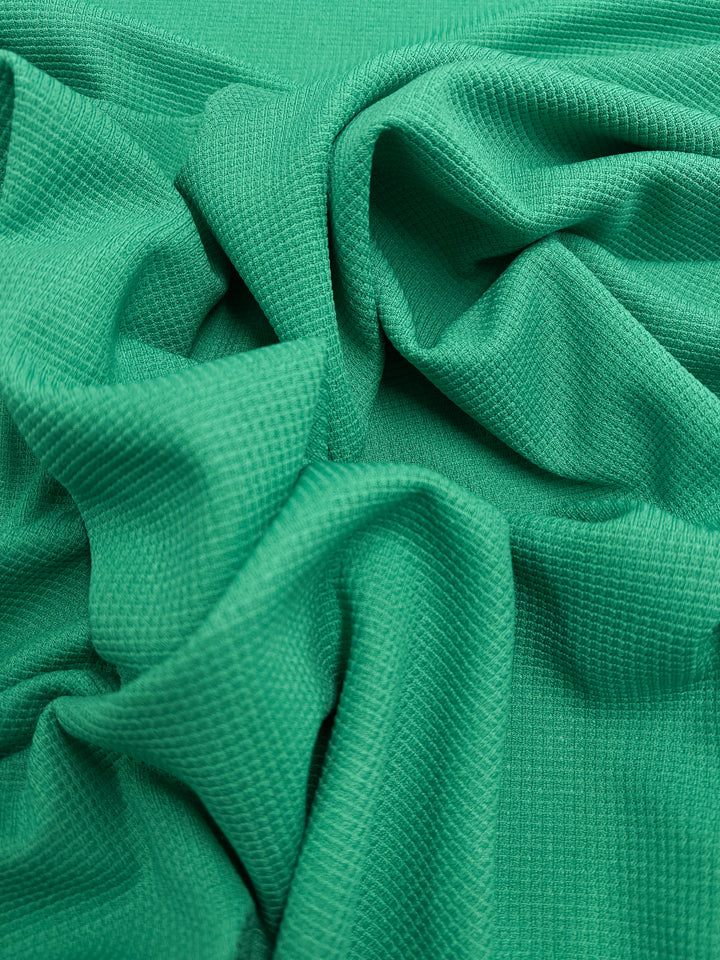 A close-up photo of a textured green waffle fabric with a wavy pattern. The material appears soft and has a grid-like texture. The folds and creases create a dynamic, undulating surface. This is the Waffle Knit - Jade Cream - 170cm by Super Cheap Fabrics.