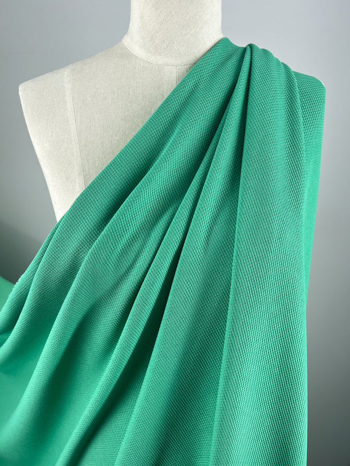 A mannequin draped in a bright teal, textured fabric stands gracefully. The fabric has a slight texture and appears to be soft and flowing, reminiscent of Waffle Knit - Jade Cream - 170cm from Super Cheap Fabrics. The background sets a simple yet elegant scene with its plain, light gray color.