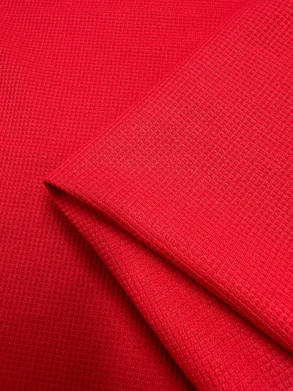 A close-up image of bright red Super Cheap Fabrics Waffle Knit - Red Alert - 170cm with a noticeable, three-dimensional appearance. The fabric is folded over itself, showcasing the intricate pattern and color variations created by the folds.